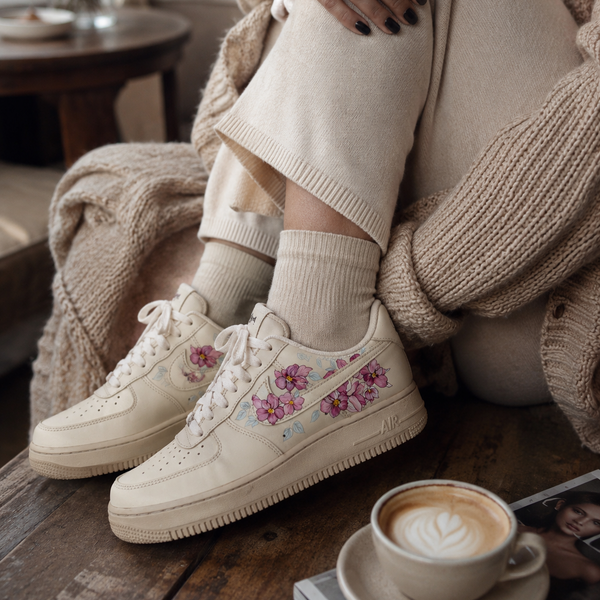 A person seated indoors wearing coffee dyed sneakers with orchids painted onto them.