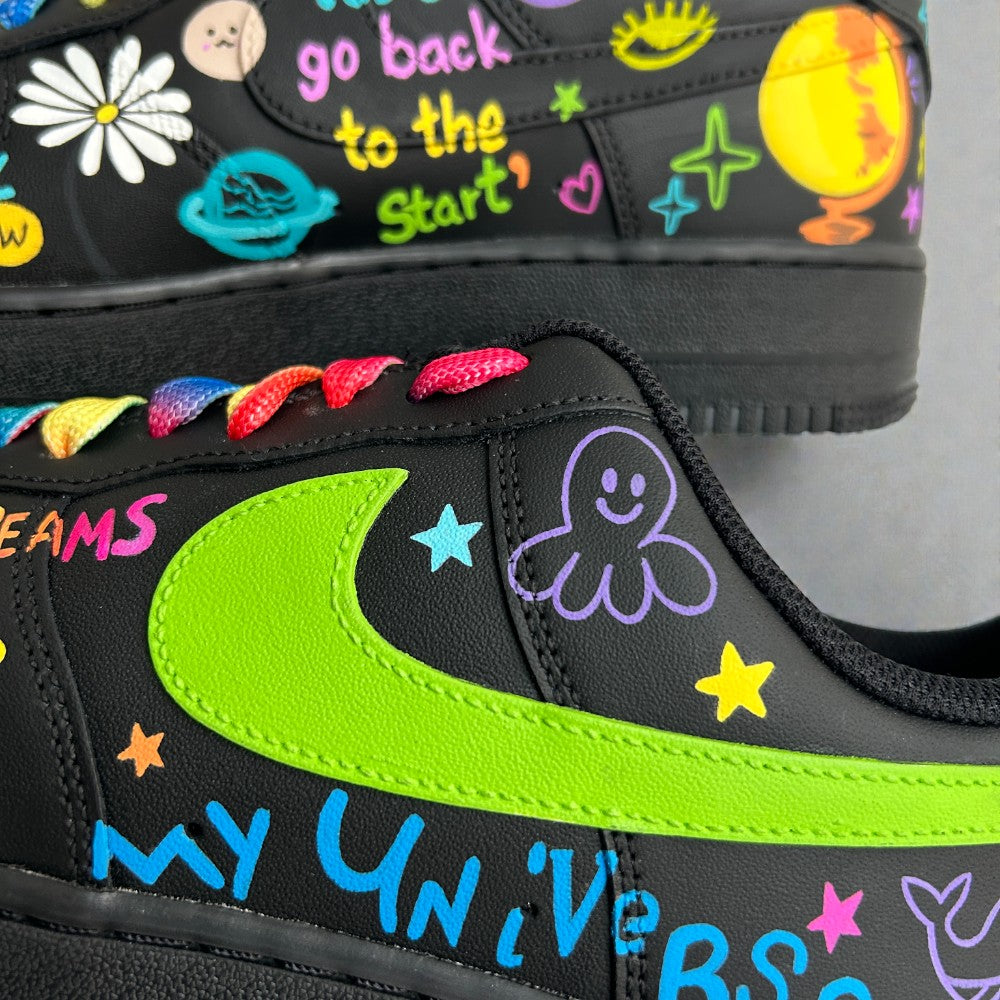 Close-up of black custom Nike Air Force 1 sneakers with neon green swoosh, colourful hand-painted doodles, rainbow laces, stars, moon, and lyric-style text reading “my universe” and “go back to the start”.