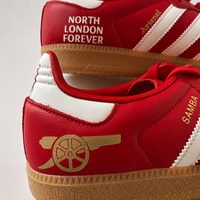 A pair of red low-top Adidas red samba sneakers featuring Arsenal icons. 