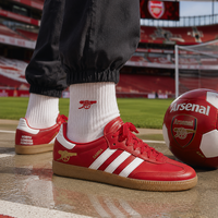 Red Arsenal custom Adidas sambas with white stripes and gold accents, worn with white Arsenal socks on a wet stadium track with a red Arsenal soccer ball nearby.