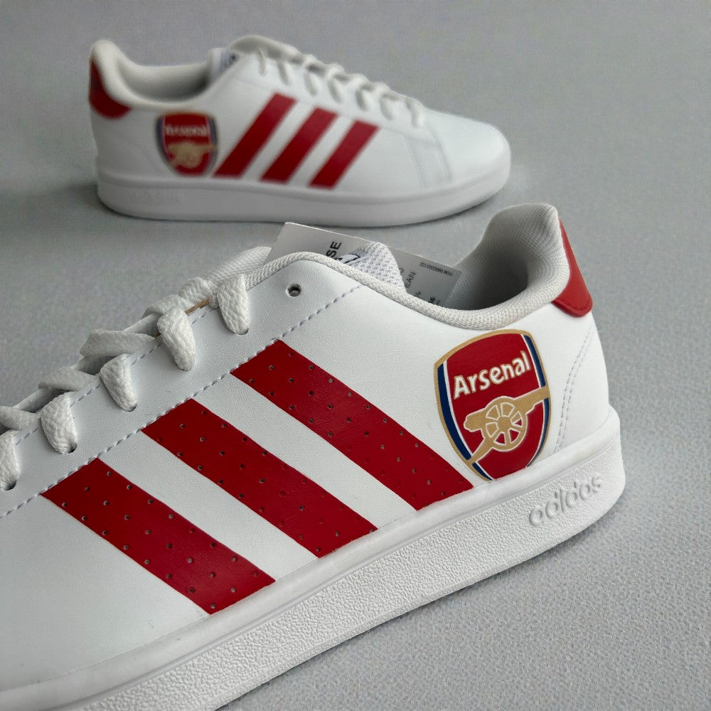 Close-up of custom Adidas sneakers featuring the Arsenal FC crest and red three-stripe detailing on a white base.