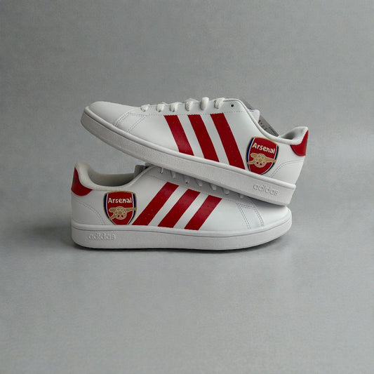 Custom Adidas sneakers inspired by Arsenal Football Club, featuring the iconic Arsenal crest on the heel, bold red three-stripe detailing, and a clean white leather base, photographed in a stacked side profile.