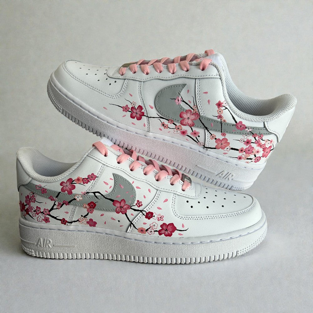 Custom hand-painted Nike Air Force 1 sneakers featuring delicate cherry blossom artwork inspired by Japanese sakura, soft pink laces, and floral branch detailing painted by hand on a white leather base, photographed in a stacked side profile.