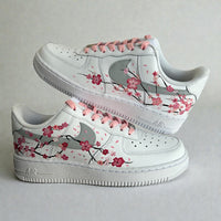 Custom hand-painted Nike Air Force 1 sneakers featuring delicate cherry blossom artwork inspired by Japanese sakura, soft pink laces, and floral branch detailing painted by hand on a white leather base, photographed in a stacked side profile.