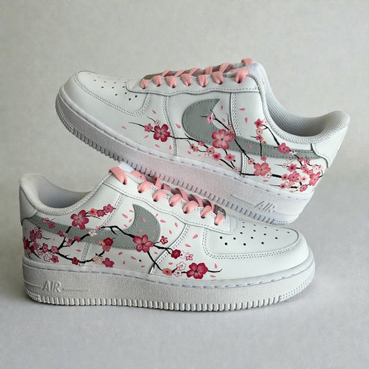 Custom hand-painted Nike Air Force 1 sneakers featuring delicate cherry blossom artwork inspired by Japanese sakura, soft pink laces, and floral branch detailing painted by hand on a white leather base, photographed in a stacked side profile.