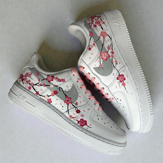 Custom hand-painted Nike Air Force 1 sneakers featuring delicate cherry blossom artwork inspired by Japanese sakura, flowing floral branches, soft pink laces, and hand-painted detailing on a white leather base, photographed as an angled pair on a clean neutral background.