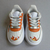 Top view of custom hand-painted kids Nike Air Force 1 sneakers featuring orange leather accents, flame artwork on the toe box, white perforated leather panels, and classic lace-up detailing.