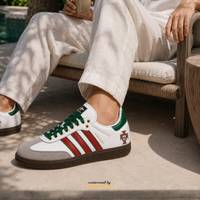 A person seated on a cushioned outdoor chair wearing a white low-top Adidas Samba sneaker with green laces, green heel tab, red side stripes, a small Portugal crest, and a brown gum sole. The photo is taken in daylight on a patio.