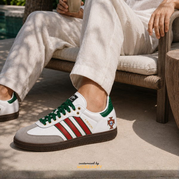 A person seated on a cushioned outdoor chair wearing a white low-top Adidas Samba sneaker with green laces, green heel tab, red side stripes, a small Portugal crest, and a brown gum sole. The photo is taken in daylight on a patio.