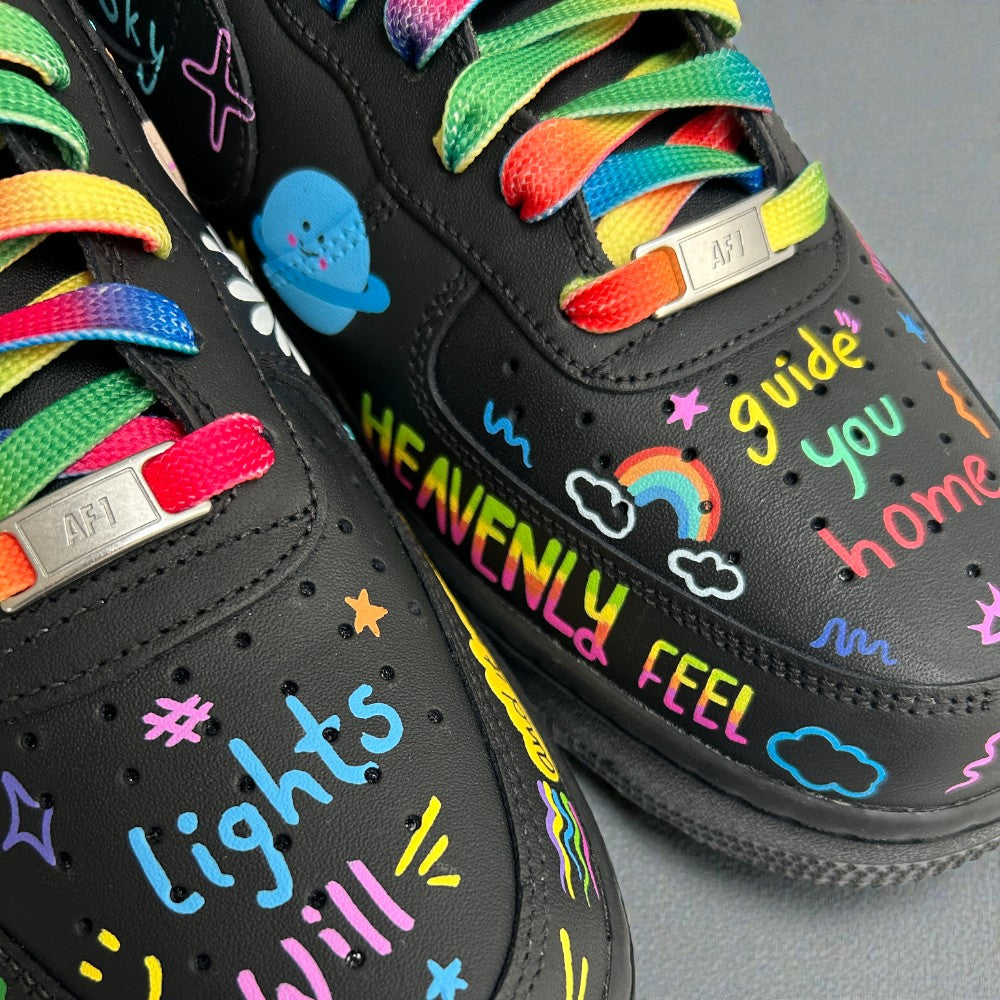 Close-up of black custom Nike Air Force 1 sneakers with rainbow laces, hand-painted doodles, clouds, stars, and colourful text reading “heavenly feel” and “guide you home”.