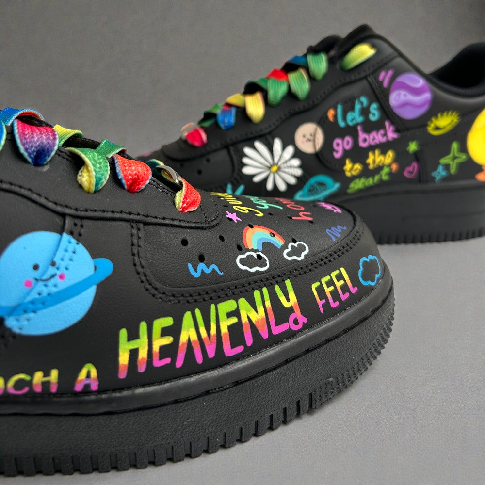 Close-up of black custom Nike Air Force 1 sneakers with rainbow laces, hand-painted doodles, planets, flowers, and colourful text reading “Heavenly Feel”.