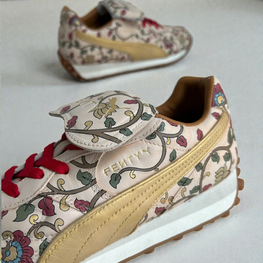 Close-up detail of custom hand-painted Puma sneakers featuring intricate floral artwork inspired by Indian motifs, metallic gold side panel, red laces, and premium leather detailing, photographed on a clean neutral background.