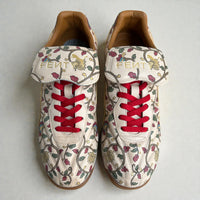 Top-down view of custom hand-painted Puma sneakers featuring intricate floral artwork inspired by Indian motifs, red laces, gold FENTY tongue branding, and premium leather detailing, photographed on a clean neutral background.