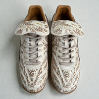 Top-down view of custom hand-painted Puma sneakers featuring intricate tribal-inspired artwork in earthy beige and brown tones, premium leather finish, and gold FENTY tongue branding, photographed on a clean neutral background.