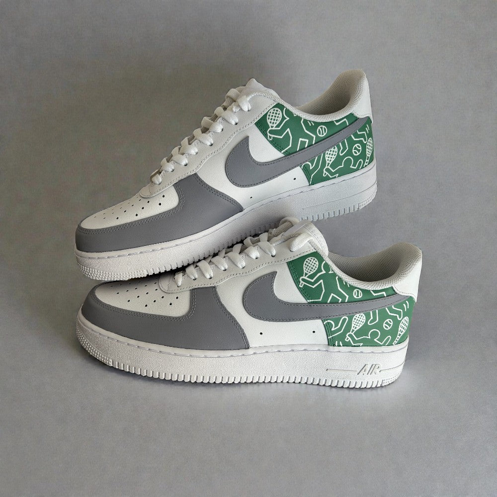 Side view of custom Nike Air Force 1 sneakers featuring grey and white leather panels, a green hand-illustrated heel design with sports icons, white laces, and the classic AF1 silhouette.