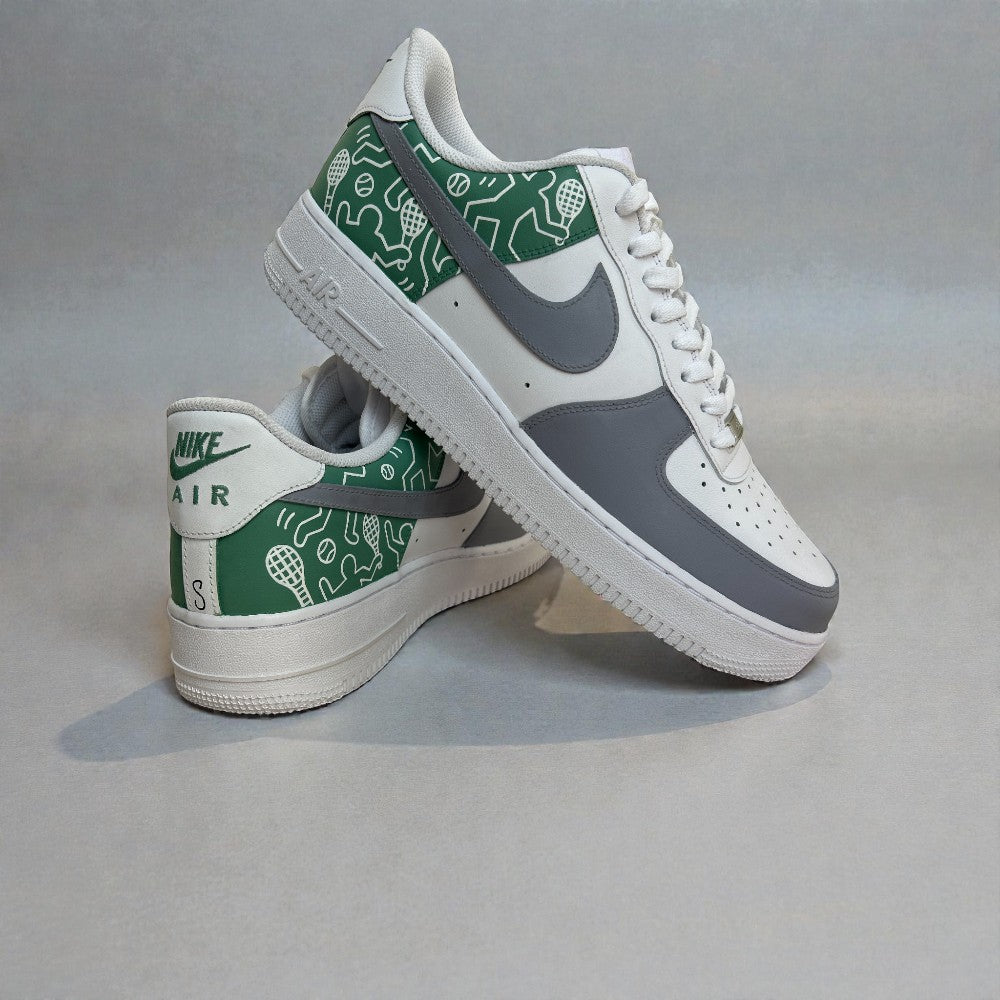 Angled side view of custom Nike Air Force 1 sneakers featuring grey and white leather panels, green hand-illustrated heel artwork with sports icons, white laces, and the classic AF1 silhouette.