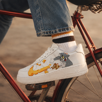 A white Nike Air Force 1 sneaker with a custom cartoon graphic of Tom and Jerry, shown on a person's foot while sitting on a red bicycle.
